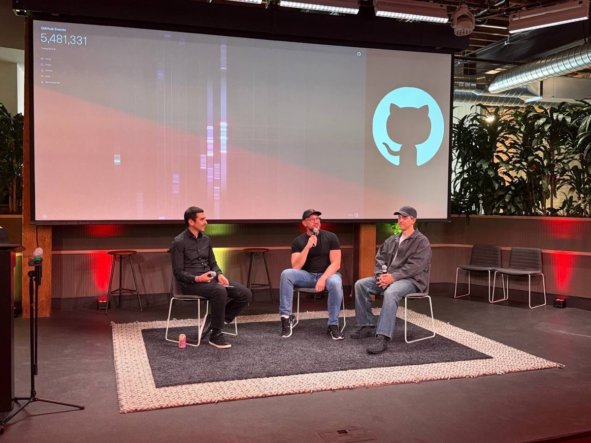Three people seated on stage mid-conversation at GitHub HQ, with the Octocat logo and a live GitHub-events counter glowing on the big screen behind them.